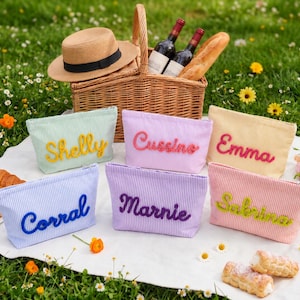 May include: Six rectangular zipped pouches made of striped fabric in pastel colours, each featuring a name embroidered in a contrasting colour. The names are Shelly, Cussino, Emma, Corral, Marnie, and Sabrina. The pouches are arranged on a white cloth.
