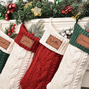 Op de afbeelding: Vier gepersonaliseerde kerstsokken in groen, rood en cr&egrave;me, elk met een leren naamlabel. De sokken zijn kabelgebreid en versierd met feestelijk groen en ornamenten, perfect voor de feestdagen.