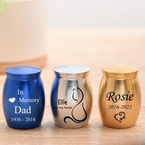 以下が含まれることがあります： 3つの小さな金属製のペット用骨壺。青い骨壺には「In Memory Dad 1936-2018」と印刷されています。銀色の骨壺には猫のシルエットと「Ellie Love You Forever」が描かれています。金色の骨壺には「Rosie 2016-2022」と書かれています。