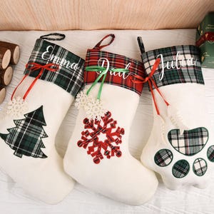 May include: Three white Christmas stockings with plaid cuffs and decorative accents. Each stocking is personalized with a name: "Emma", "Dial", and "Julia". The stockings feature a Christmas tree, snowflake, and paw print design.