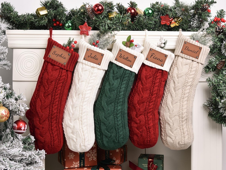 Op de afbeelding: Vijf kabelgebreide kerstsokken in rood, wit, groen en beige hangen aan een witte schoorsteenmantel. Elke sok heeft een leren naamlabel. Feestelijk groen en ornamenten versieren de schoorsteenmantel.