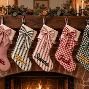 Puede incluir: Cinco calcetines de Navidad colgados en fila, cada uno con un puño de color crema con borde con volantes y un gran lazo de tela. Los calcetines muestran los nombres Olivia, Ethan, Sienna, Jack y Harper en letras bordadas.