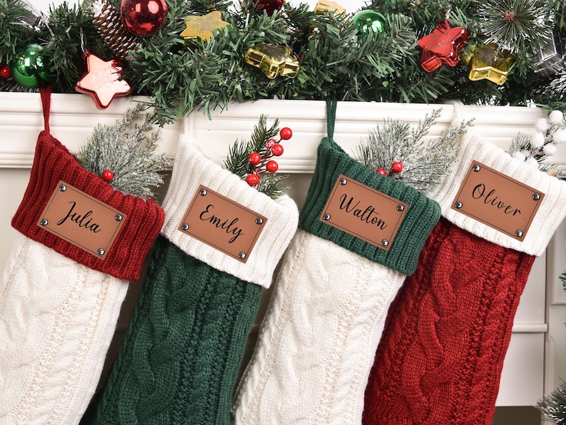 Op de afbeelding: Vier gepersonaliseerde kerstsokken in rood, groen en wit kabelbreisel. Elke sok heeft een bruin leren label met een naam: Julia, Emily, Walton en Oliver. Feestelijke vakantiedecoratie.