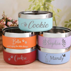Peut inclure: Empilement de cinq gamelles pour animaux de compagnie en acier inoxydable. Les bols sont de couleurs variées : bleu clair, orange, rouge, lavande et bleu clair. Chaque bol a un bord argenté et une bande colorée avec un nom d'animal.