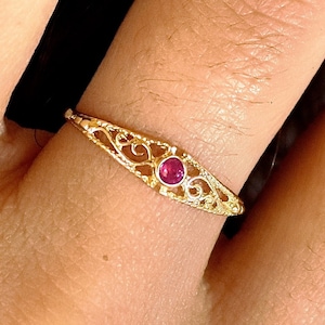 Könnte beinhalten: Ein goldener Ring mit einem zentralen, runden, rosafarbenen Edelstein, der in ein zartes, längliches Filigran-Metallband eingefasst ist. Das komplexe durchbrochene Design erstreckt sich über die Oberseite des Bandes und zeigt ein detailliertes Muster.