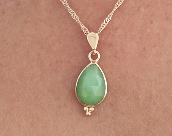 May include: A gold-coloured necklace featuring a light green, teardrop-shaped pendant. The pendant is set in gold and has small decorative elements. The chain is a delicate, twisted design.