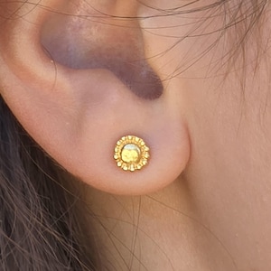 May include: A close-up of a gold-colored stud earring with a floral design. The earring is round with a textured border and a smooth center. The earring is worn on an ear.