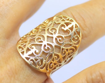Könnte beinhalten: Ein goldfarbener Ring mit einem durchbrochenen Design aus Wirbeln und geometrischen Mustern. Der Ring hat eine große, längliche Oberfläche. Der Ring wird an einem Finger getragen. Das Design des Rings ist auf der Haut sichtbar.