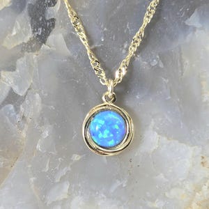 May include: A gold-toned necklace featuring a round pendant with a vibrant blue opal stone. The pendant is encircled by a delicate, twisted gold frame, and hangs from a matching gold chain. The necklace is displayed against a neutral background.