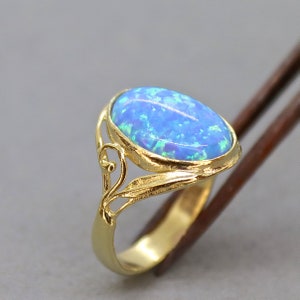 May include: A gold ring featuring an oval, blue opal gemstone. The ring has a decorative filigree design on the sides. The opal displays a vibrant play of color, with hints of green and blue. The ring is set against a neutral background.