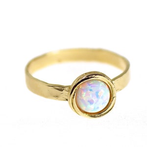 Könnte beinhalten: Ein goldener Ring mit einem weißen Opal-Edelstein, der in einer Zargenfassung gefasst ist. Der Ringarm ist texturiert und hat eine gehämmerte Oberfläche.