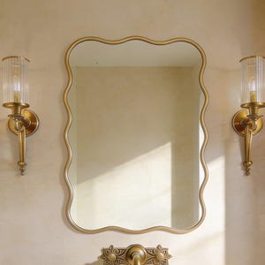 Peut inclure: Ensemble de vanité de salle de bain avec un miroir doré à bord ondulé, un évier en pierre et des appliques dorées avec des abat-jours en verre. Les luminaires ont une finition en laiton antique, créant une esthétique luxueuse et classique.