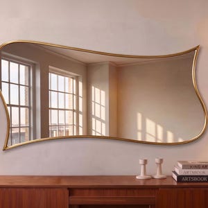 May include: A wall-mounted mirror with an irregular, wavy gold frame. The reflective surface shows a room interior with windows. The mirror is positioned above a wooden console table, flanked by two floor lamps with white shades and stacks of books.