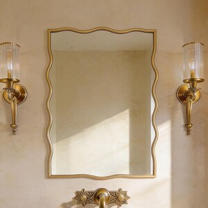 Peut inclure: Un ensemble de vanité de salle de bain avec un évier en pierre, un miroir aux bords ondulés et des appliques en laiton avec des abat-jours en verre. Le robinet et le cadre du miroir ont une finition dorée assortie. Le mur est peint en beige neutre.