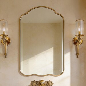 Peut inclure: Un ensemble de vanité de salle de bain avec un miroir doré à bord festonné, un évier en pierre et des appliques dorées. Le robinet présente un motif floral. Le mur est de couleur beige clair.