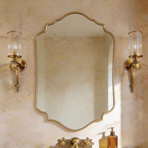 Peut inclure: Un ensemble de vanité de salle de bain avec un miroir doré à bord festonné, flanqué de deux appliques avec des abat-jours en verre. L'évier en pierre a un robinet à motif floral et un distributeur de savon.