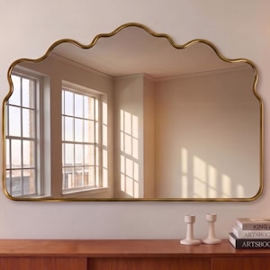 May include: A wall-mounted mirror featuring a thin, metallic gold frame with a distinct scalloped, wavy top edge. The rectangular glass surface reflects a bright room with windows, while the mirror is positioned above a wooden console table.