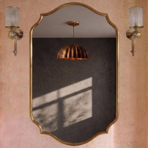 Puede incluir: Un espejo de pared con marco festoneado en tono dorado, flanqueado por dos apliques de pared verticales con pantallas de cristal estriado. El espejo refleja una pared gris oscuro, una lámpara colgante de cúpula cobriza y un grifo metálico.