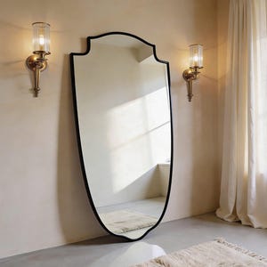 Peut inclure: Un grand miroir en forme de bouclier avec un cadre noir est monté sur un mur de couleur claire. Deux appliques avec des abat-jours en verre et des fixations en laiton encadrent le miroir. Un tapis crème est posé sur le sol.