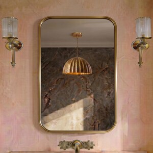 May include: A rectangular wall mirror with a thin gold frame, flanked by two vertical wall sconces featuring fluted glass shades and gold hardware. A gold pendant light with a ribbed dome shade is reflected in the mirror above a gold faucet.