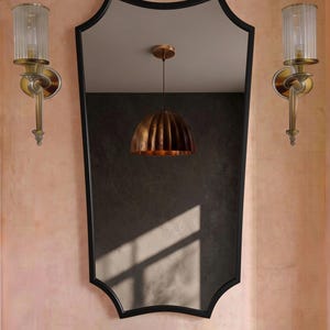 Könnte beinhalten: Ein Wandspiegel mit schwarzem Rahmen und geschwungenem Randdesign. Flankiert wird der Spiegel von zwei Wandleuchten aus Messing mit gerippten Glasschirmen. Unter dem Spiegel befindet sich eine Messingarmatur mit floralen Zierplatten.