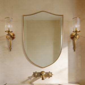 Puede incluir: Un conjunto de tocador de baño con un espejo en forma de escudo con marco dorado, dos apliques con pantallas de vidrio, un lavabo de piedra y un grifo dorado. La pared tiene un acabado beige claro texturizado. Diseño elegante.