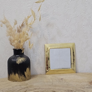 Peut inclure: Un vase noir et doré avec des fleurs séchées et un petit miroir carré doré sur une surface en bois. Le vase présente un motif texturé et nervuré avec des coups de peinture dorés. Le miroir a un cadre doré.