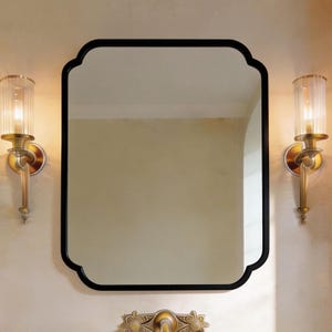 Peut inclure: Un miroir rectangulaire à cadre noir avec un bord supérieur festonné est suspendu au-dessus d'un robinet doré. Deux appliques murales avec des abat-jours en verre cannelé transparent et des fixations en laiton encadrent le miroir.