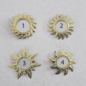 Peut inclure: Quatre miroirs dorés en forme de soleil, chacun avec un centre blanc et un numéro noir de 1 à 4. Les miroirs ont un motif texturé en forme de feuille sur les bords. Montés sur un mur blanc cassé texturé.