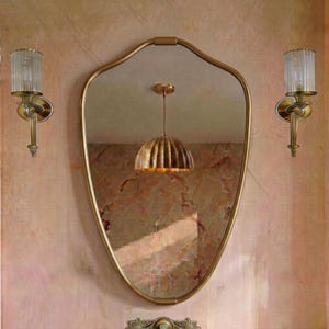Puede incluir: Un espejo de pared con forma de escudo y un marco dorado fino, flanqueado por dos apliques de pared a juego con pantallas de cristal estriado vertical y herrajes dorados. El espejo refleja una lámpara colgante con una pantalla dorada plisada.