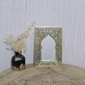 Peut inclure: Un miroir décoratif avec un cadre doré et un motif de filigrane noir. Le miroir a un sommet en forme d'arche et est exposé sur une surface en bois. Un petit vase noir et or avec des fleurs séchées est à côté du miroir.