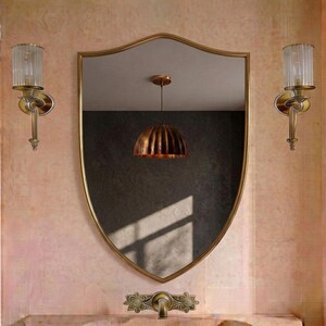 Peut inclure: Un miroir mural en forme de bouclier avec un cadre doré fin, flanqué de deux appliques murales dorées assorties avec des abat-jours en verre cannelé. Un robinet doré aux accents floraux décoratifs est monté en dessous, reflétant une suspension.