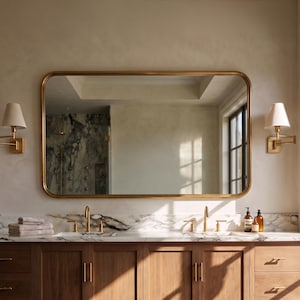 A wide horizontal rectangle solid brass mirror with smoothly rounded corners, mounted beautifully above a dual-sink vanity in a modern master bathroom setting.