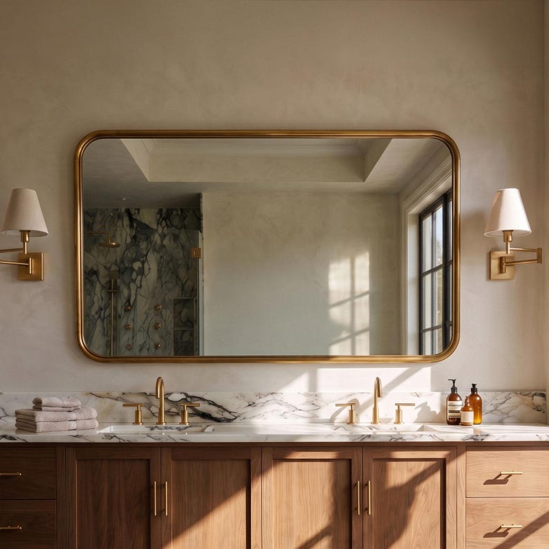 A wide horizontal rectangle solid brass mirror with smoothly rounded corners, mounted beautifully above a dual-sink vanity in a modern master bathroom setting.