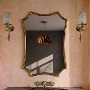 May include: A wall-mounted mirror with a gold-toned, scalloped frame. Flanking the mirror are two matching gold-toned wall sconces with fluted glass shades. A gold-toned metal faucet is positioned below the mirror on a dark countertop surface.