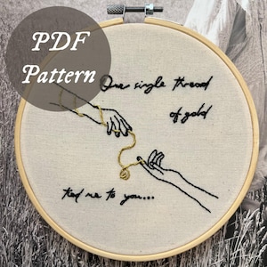 May include: A PDF pattern for embroidery art. The design features two hands reaching for each other, connected by a gold chain. The text reads "One single thread of gold" and "tied me to you..." on a cream-colored fabric within a wooden hoop.