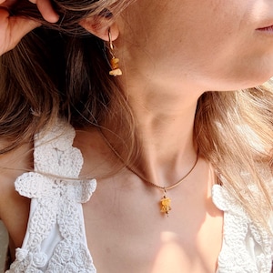 May include: A gold-toned metal necklace featuring a small, irregular yellow gemstone pendant on a delicate chain, paired with a matching gold-toned hoop earring that holds a single, small, irregular yellow gemstone charm.