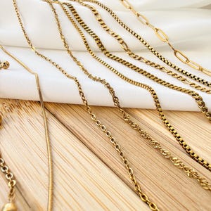 May include: A collection of gold-toned metal chains featuring various link styles, including a bead chain, a snake chain, a twisted rope chain, a box chain, a curb chain, a Figaro chain, and a long oval link chain arranged in a parallel row.