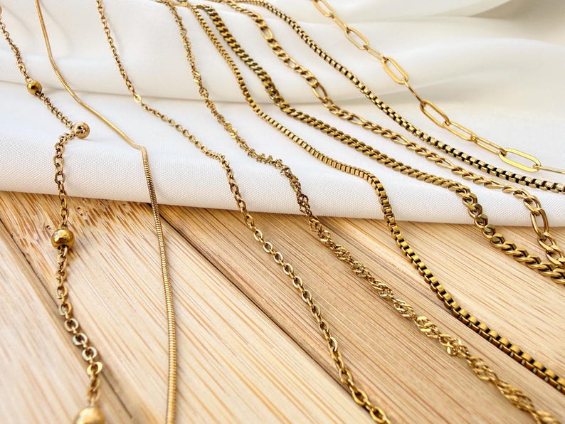 May include: A collection of gold-toned metal chains featuring various link styles, including a bead chain, a snake chain, a twisted rope chain, a box chain, a curb chain, a Figaro chain, and a long oval link chain arranged in a parallel row.