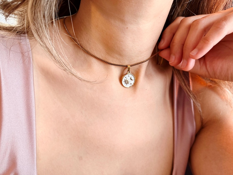 May include: A gold-toned chain necklace featuring a small, round pendant. The pendant has a white enamel background with a gold-colored sun, moon, and star design engraved on the surface. The necklace rests against the neck on a thin metal chain.