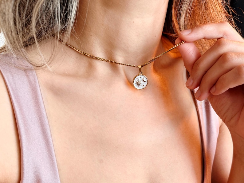 May include: A gold-toned chain necklace featuring a circular pendant with a white background. The pendant displays a gold sun and moon motif with small star accents. The necklace rests against the collarbone, showcasing a delicate link design.