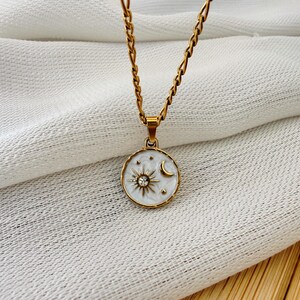 May include: A gold-toned chain necklace featuring a round pendant with a white enamel background. The pendant displays a gold sun motif with a central clear crystal, a gold crescent moon, and small gold stars engraved on the surface.