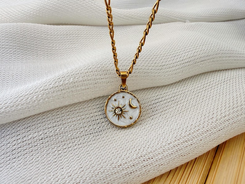 May include: A gold-toned chain necklace featuring a round pendant with a white enamel background. The pendant displays a gold sun motif with a central clear crystal, a gold crescent moon, and small gold stars engraved on the surface.
