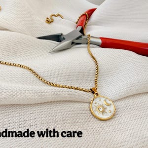 May include: A gold-toned box chain necklace featuring an oval pendant with a white enamel inlay, a gold sun and moon motif, and a central clear stone. The necklace is accompanied by red-handled metal pliers. The text reads: Handmade with care.