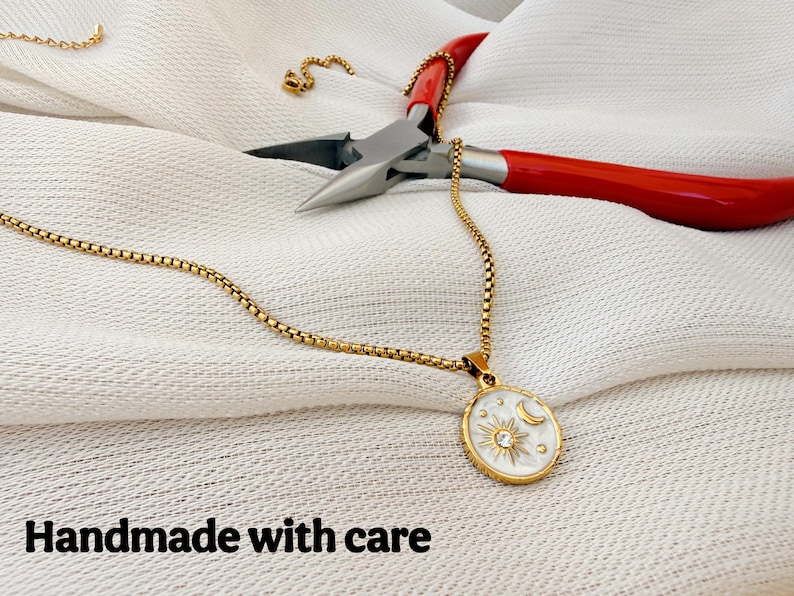 May include: A gold-toned box chain necklace featuring an oval pendant with a white enamel inlay, a gold sun and moon motif, and a central clear stone. The necklace is accompanied by red-handled metal pliers. The text reads: Handmade with care.
