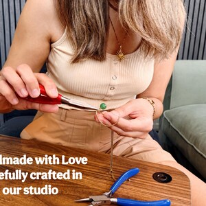 May include: A gold chain necklace featuring a small green spherical bead, being assembled with red-handled needle-nose pliers. The text 'Handmade with Love Carefully crafted in our studio' is visible. A second pair of blue-handled pliers rests nearby.