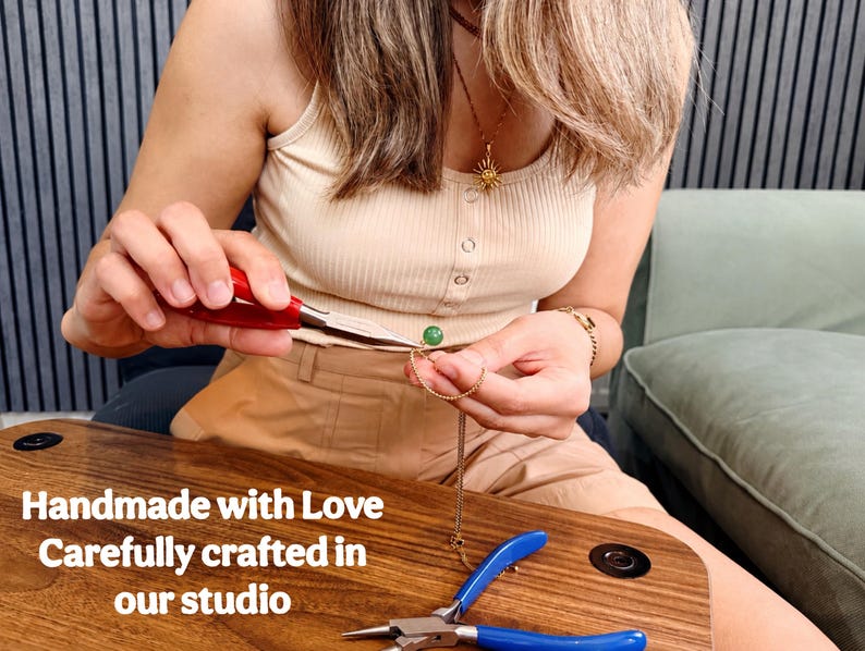 May include: A gold chain necklace featuring a small green spherical bead, being assembled with red-handled needle-nose pliers. The text 'Handmade with Love Carefully crafted in our studio' is visible. A second pair of blue-handled pliers rests nearby.