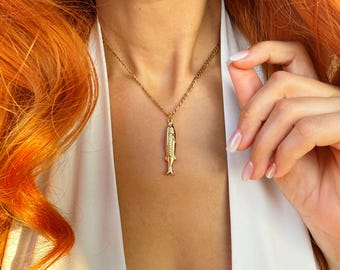 May include: A gold-toned necklace featuring a pendant shaped like a small fish. The pendant hangs from a delicate gold-colored chain. The fish design includes detailed scale textures and a defined tail fin, creating a simple and polished accessory.