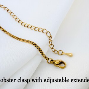 May include: A gold-toned box chain necklace featuring a lobster clasp and an adjustable extender chain. The extender chain ends with a small, smooth teardrop-shaped charm. The text "Lobster clasp with adjustable extender" is printed below.
