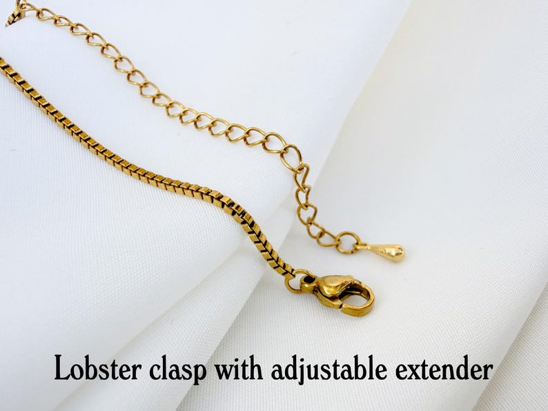 May include: A gold-toned box chain necklace featuring a lobster clasp and an adjustable extender chain. The extender chain ends with a small, smooth teardrop-shaped charm. The text "Lobster clasp with adjustable extender" is printed below.